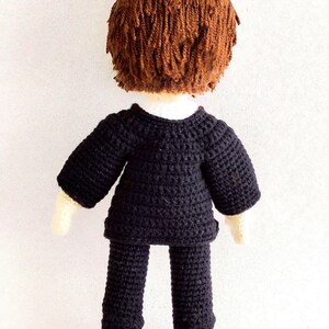 May include: A crocheted doll wearing a black suit with brown hair. The doll is standing on a white surface.