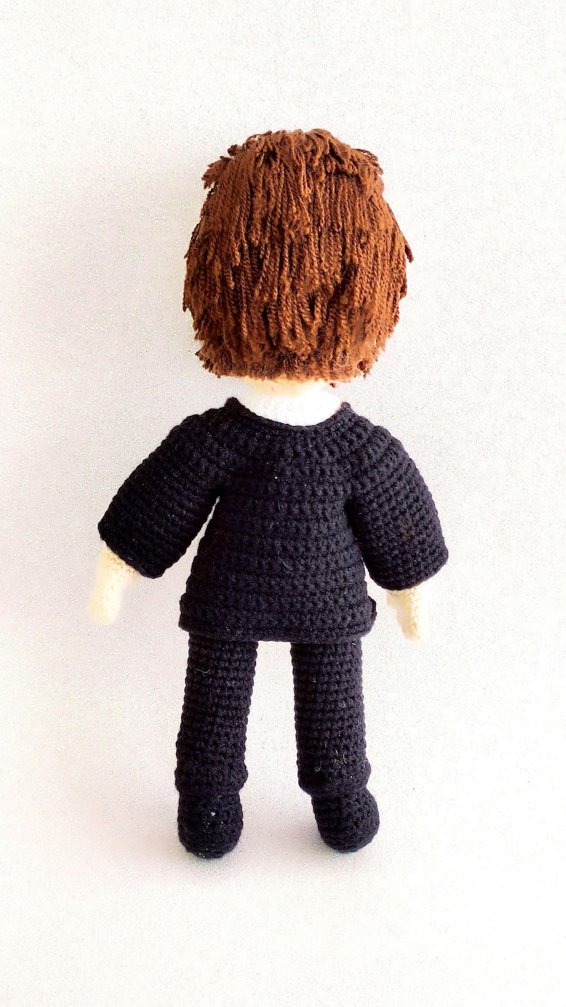 May include: A crocheted doll wearing a black suit with brown hair. The doll is standing on a white surface.