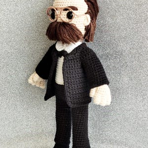 May include: A crocheted plush toy of a person wearing a black suit, white shirt, and bow tie. The toy has brown hair, a brown moustache, and round glasses.