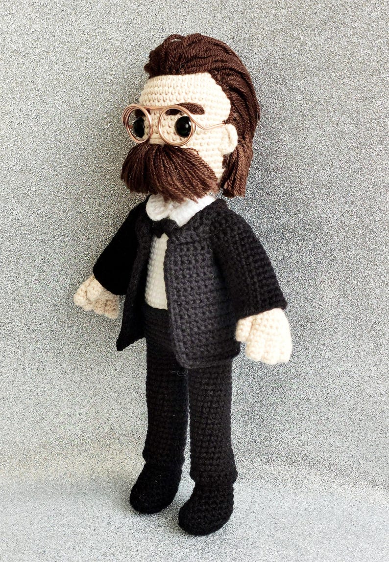 May include: A crocheted plush toy of a person wearing a black suit, white shirt, and bow tie. The toy has brown hair, a brown moustache, and round glasses.