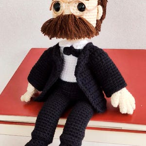 May include: Crocheted doll wearing a black suit with a white shirt and bow tie. The doll has brown hair, a brown moustache, and is wearing round glasses.