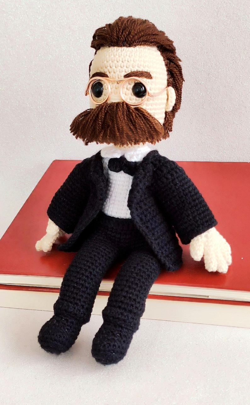 May include: Crocheted doll wearing a black suit with a white shirt and bow tie. The doll has brown hair, a brown moustache, and is wearing round glasses.