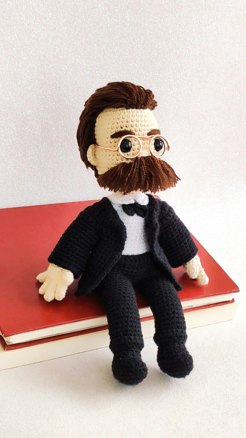 May include: Crocheted doll wearing a black suit, white shirt, and bow tie. The doll has brown hair, a brown moustache, and is wearing round glasses.