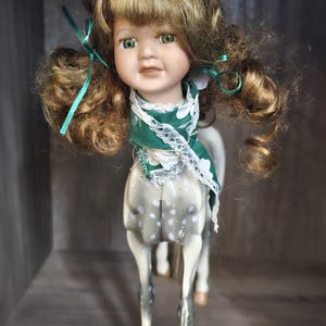 May include: A porcelain doll with a human face and a horse body. The doll has green eyes, brown hair, and a green scarf with white lace. The horse body is gray with white spots and has four legs.
