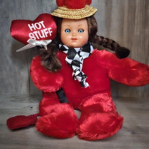 May include: A vintage doll with blue eyes, wearing a red fuzzy outfit, a straw hat with red pom-poms, and a black and white checkered scarf. The doll holds a red heart-shaped sign that reads "HOT STUFF".