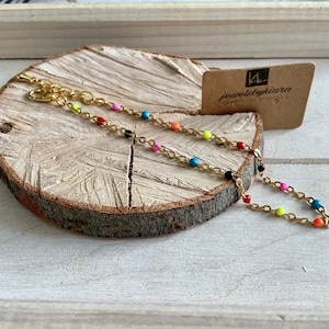 May include: A gold chain bracelet with colorful beads. The beads are in shades of red, pink, blue, green, and yellow. The bracelet is on a wooden surface with a small tag that says "jewelsbykiara".