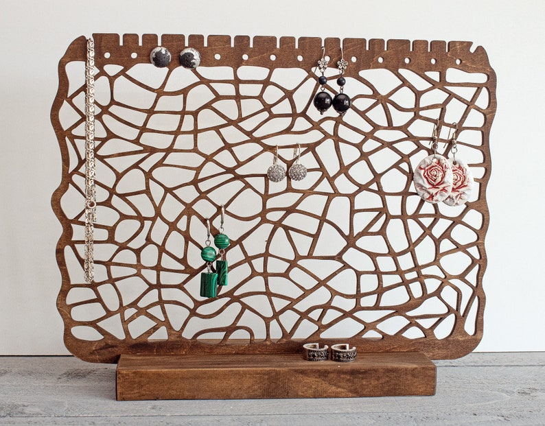 May include: Wooden jewellery stand with a delicate, intricate design. It features a variety of earrings, including black, green, and floral designs. The stand also has a small shelf for rings.
