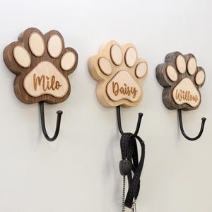 Op de afbeelding: Drie houten wandhaken in de vorm van een pootafdruk met zwarte metalen haken. Elke pootafdruk heeft een andere kleur en een naam: Milo, Daisy en Willow. Een zwarte en zilveren hondenriem hangt aan de middelste haak. De haken zijn aan een witte muur bevestigd.