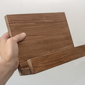K&ouml;nnte beinhalten: Ein brauner Holz-Kochbuchst&auml;nder mit einer schr&auml;gen Oberfl&auml;che zum Halten eines Kochbuchs oder Tablets. Der St&auml;nder ist aus Naturholz mit rustikalem Finish.