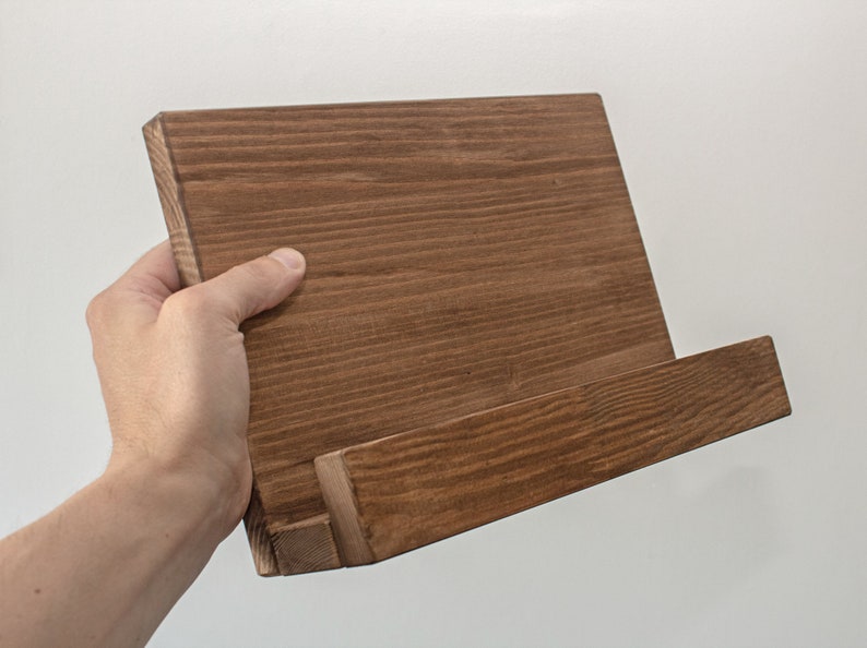 K&ouml;nnte beinhalten: Ein brauner Holz-Kochbuchst&auml;nder mit einer schr&auml;gen Oberfl&auml;che zum Halten eines Kochbuchs oder Tablets. Der St&auml;nder ist aus Naturholz mit rustikalem Finish.