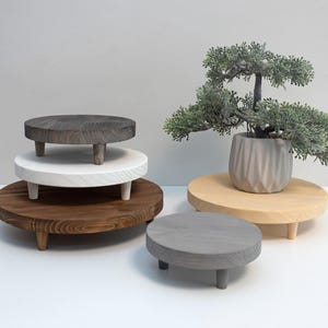 Könnte beinhalten: Eine Sammlung von Pflanzenständern aus Holz in verschiedenen Größen und Ausführungen. Die Ständer sind rund mit kurzen, konischen Beinen. Die Ausführungen umfassen Dunkelgrau, Weiß und Braun. Ein kleiner Bonsai im Topf steht auf einem Naturholzständer.
