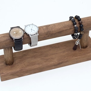 May include: A wooden watch and bracelet stand with two watches and a beaded bracelet with a tassel. The stand is brown and has a rectangular base.