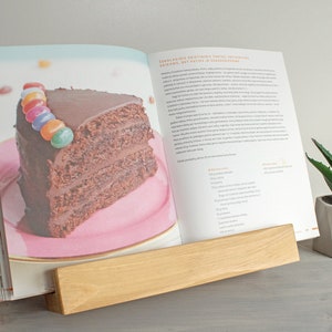 K&ouml;nnte beinhalten: Ein Holzbuchst&auml;nder h&auml;lt ein Kochbuch offen auf einem Rezept f&uuml;r Schokoladenkuchen mit einem St&uuml;ck Kuchen auf einem rosa Teller. Der Kuchen ist mit bunten S&uuml;&szlig;igkeiten verziert. Der Buchst&auml;nder steht auf einer grauen Oberfl&auml;che neben einer kleinen Sukkulente im Topf.