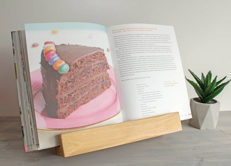 K&ouml;nnte beinhalten: Ein Holzbuchst&auml;nder h&auml;lt ein Kochbuch offen auf einem Rezept f&uuml;r Schokoladenkuchen mit einem St&uuml;ck Kuchen auf einem rosa Teller. Der Kuchen ist mit bunten S&uuml;&szlig;igkeiten verziert. Der Buchst&auml;nder steht auf einer grauen Oberfl&auml;che neben einer kleinen Sukkulente im Topf.
