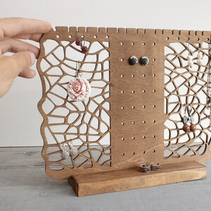 May include: Wooden earring and necklace stand with a geometric design. The stand has a brown finish and features multiple holes for hanging earrings and a small shelf for necklaces.