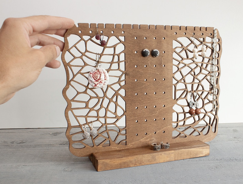 May include: Wooden earring and necklace stand with a geometric design. The stand has a brown finish and features multiple holes for hanging earrings and a small shelf for necklaces.