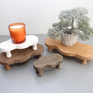 Puede incluir: Conjunto de elevadores de madera en varios tamaños y acabados. Uno sostiene una vela naranja en un tarro de cristal, otro exhibe un pequeño bonsái en una maceta blanca. Los elevadores están teñidos en blanco, marrón y gris.