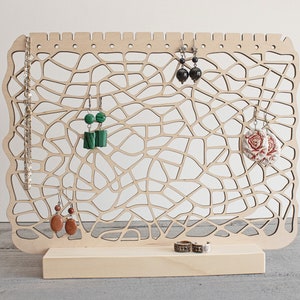 May include: Wooden earring stand with a geometric design. The stand has a base and a vertical panel with multiple holes for hanging earrings. There are earrings with green, black, and floral designs hanging on the stand.