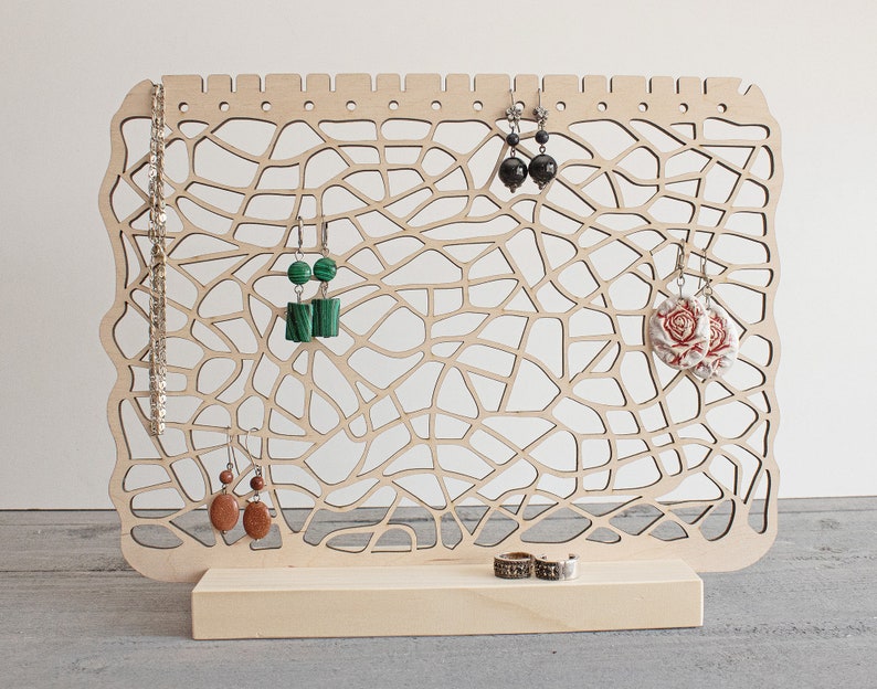 May include: Wooden earring stand with a geometric design. The stand has a base and a vertical panel with multiple holes for hanging earrings. There are earrings with green, black, and floral designs hanging on the stand.