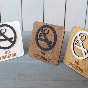 Può includere: Tre cartelli in legno con la scritta "No Smoking" e un'immagine stilizzata di una sigaretta con una linea diagonale che la attraversa. I cartelli sono di colori diversi: bianco, marrone e marrone chiaro.