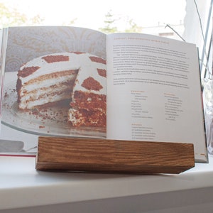 K&ouml;nnte beinhalten: Ein Holzbuchst&auml;nder mit einem ge&ouml;ffneten Kochbuch darauf. Das Kochbuch enth&auml;lt ein Rezept f&uuml;r einen mehrschichtigen Kuchen mit Schokoladen- und Sahnef&uuml;llung. Der Kuchen befindet sich auf einem wei&szlig;en Teller.