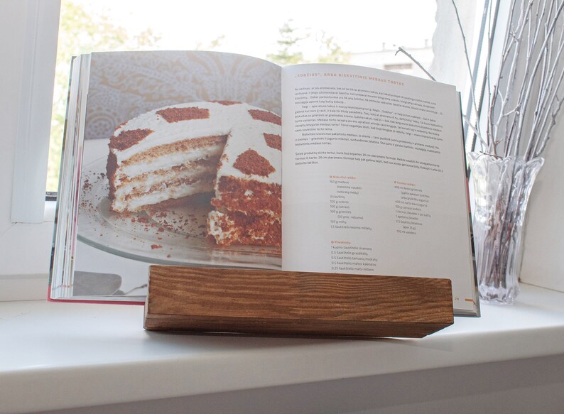 K&ouml;nnte beinhalten: Ein Holzbuchst&auml;nder mit einem ge&ouml;ffneten Kochbuch darauf. Das Kochbuch enth&auml;lt ein Rezept f&uuml;r einen mehrschichtigen Kuchen mit Schokoladen- und Sahnef&uuml;llung. Der Kuchen befindet sich auf einem wei&szlig;en Teller.