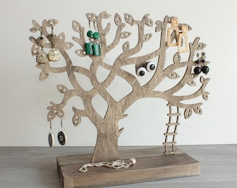 Puede incluir: Soporte para joyas de madera en forma de árbol con una escalera y múltiples ramas para colgar pendientes y un collar. El soporte tiene un acabado de madera marrón.