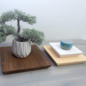 Op de afbeelding: Een decoratieve display met een kleine bonsai-boom in een witte pot op een vierkante houten dienblad. Een stapel van twee vierkante houten trays, één wit en één licht hout, bevat een kleine, ronde, blauw en goudkleurig patroon sieradendoosje.