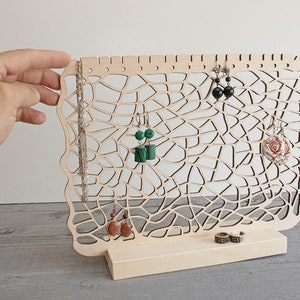 May include: A wooden jewellery stand with a delicate, intricate design. The stand features multiple hooks for earrings and a small shelf for rings. The stand is made of natural wood and has a light brown finish.