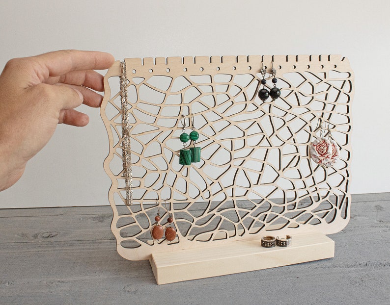May include: A wooden jewellery stand with a delicate, intricate design. The stand features multiple hooks for earrings and a small shelf for rings. The stand is made of natural wood and has a light brown finish.