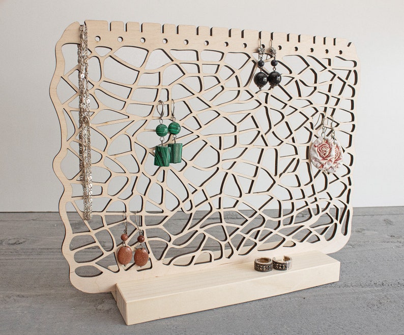 May include: Wooden jewellery stand with a delicate, intricate design. The stand features a large, open area for hanging earrings and a smaller base for rings or bracelets. The stand is made of light-coloured wood and has a natural finish.