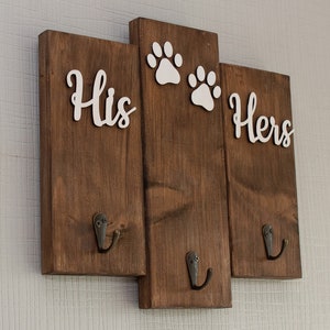 May include: A wooden wall-mounted key holder with two hooks on each side. The middle section has two white paw prints. The left side says "His" and the right side says "Hers".
