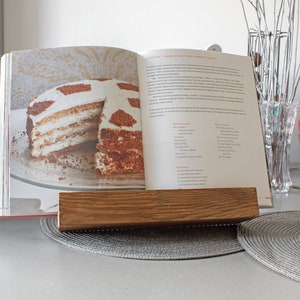 K&ouml;nnte beinhalten: Ein Holz-Kochbuchst&auml;nder mit einem Kochbuch, das auf ein Rezept f&uuml;r einen mehrschichtigen Kuchen mit wei&szlig;em Frosting und Schokoladenkr&uuml;meln ge&ouml;ffnet ist. Der Kuchen befindet sich auf einem wei&szlig;en Teller. Das Kochbuch tr&auml;gt den Titel "Anna's Delicious World Treats".