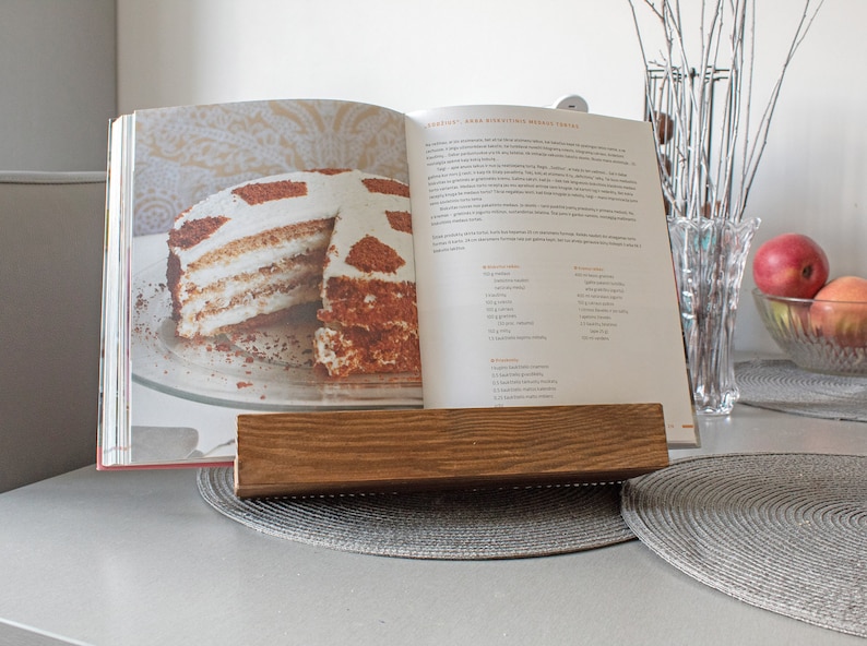 K&ouml;nnte beinhalten: Ein Holz-Kochbuchst&auml;nder mit einem Kochbuch, das auf ein Rezept f&uuml;r einen mehrschichtigen Kuchen mit wei&szlig;em Frosting und Schokoladenkr&uuml;meln ge&ouml;ffnet ist. Der Kuchen befindet sich auf einem wei&szlig;en Teller. Das Kochbuch tr&auml;gt den Titel "Anna's Delicious World Treats".