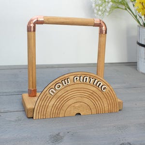 Puede incluir: Soporte de discos de madera con detalles de tubería de cobre. El soporte tiene un diseño de medio círculo con las palabras "Now Playing" y un patrón de arcoíris.