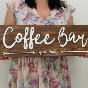 Puede incluir: Un letrero de madera marrón con letras blancas que dice "Coffee Bar" y "open daily" con flechas blancas a cada lado.