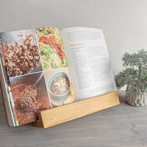 K&ouml;nnte beinhalten: Ein Holz-Kochbuchst&auml;nder mit einem Kochbuch, das auf ein Rezept f&uuml;r "Chili con carne" ge&ouml;ffnet ist. Das Kochbuch enth&auml;lt Fotos der Zutaten und des fertigen Gerichts. Der St&auml;nder steht auf einer grauen Oberfl&auml;che neben einem kleinen Topfbaum.