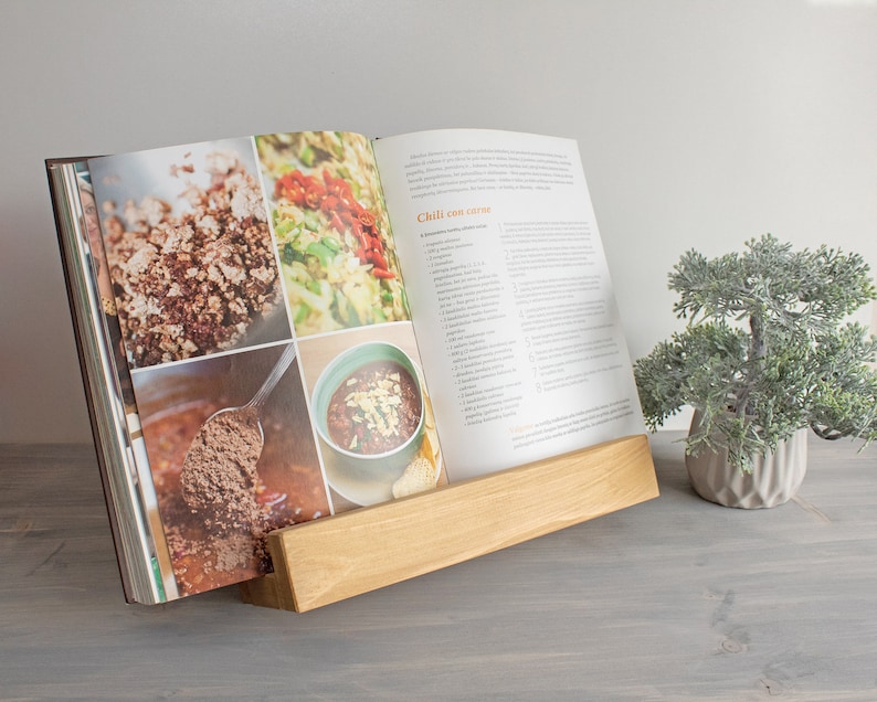 K&ouml;nnte beinhalten: Ein Holz-Kochbuchst&auml;nder mit einem Kochbuch, das auf ein Rezept f&uuml;r "Chili con carne" ge&ouml;ffnet ist. Das Kochbuch enth&auml;lt Fotos der Zutaten und des fertigen Gerichts. Der St&auml;nder steht auf einer grauen Oberfl&auml;che neben einem kleinen Topfbaum.