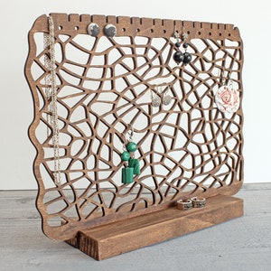 May include: Wooden jewellery stand with a web-like design. The stand has a base and a vertical section with multiple holes for hanging earrings and necklaces. The stand is brown and has a variety of jewellery hanging from it, including earrings, necklaces, and a ring.