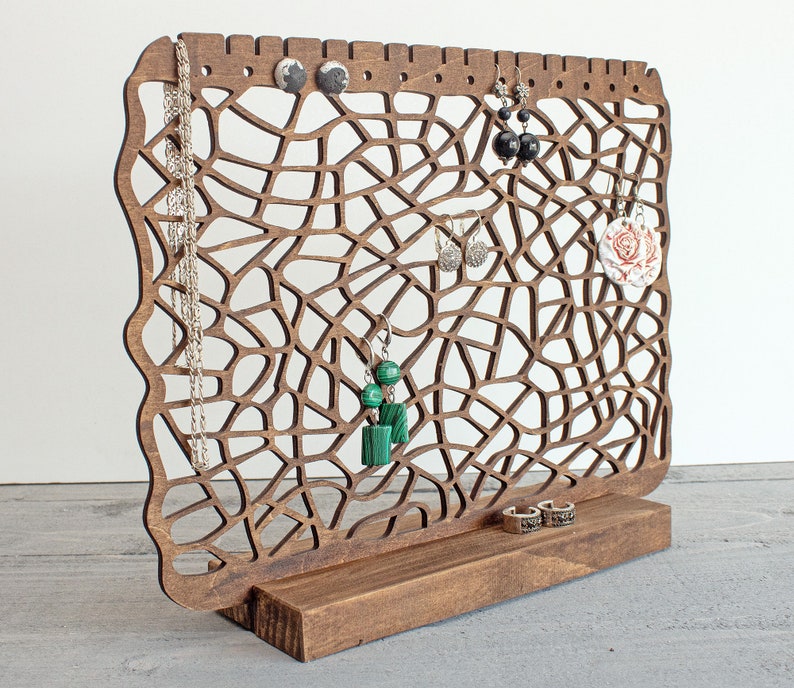 May include: Wooden jewellery stand with a web-like design. The stand has a base and a vertical section with multiple holes for hanging earrings and necklaces. The stand is brown and has a variety of jewellery hanging from it, including earrings, necklaces, and a ring.