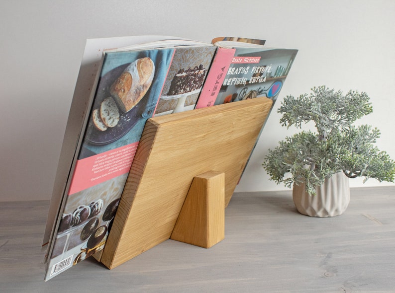 K&ouml;nnte beinhalten: Ein naturfarbener Holzst&auml;nder f&uuml;r Kochb&uuml;cher, der einen Stapel Kochb&uuml;cher mit Bildern von Speisen auf den Umschl&auml;gen h&auml;lt. Der St&auml;nder hat eine dreieckige Basis und eine rechteckige Oberseite.