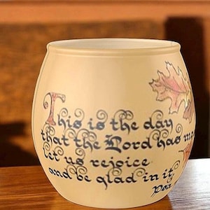 May include: A frosted glass votive candle holder featuring a rounded shape and a wide opening. The exterior is decorated with colourful autumn leaf illustrations and black calligraphy text that reads, This is the day that the Lord has made, let us rejoice and be glad in it, Psalm 118:24.