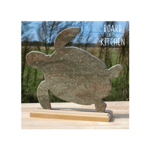 May include: A green and brown granite turtle-shaped cutting board with a wooden base. The board is standing on a wooden table. The text "BOARD IN THE KITCHEN" is printed on a white background in the upper right corner of the image.