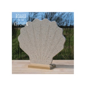 Puede incluir: Una tabla de cortar de color beige con forma de concha marina con una base de madera. La tabla tiene una superficie texturizada y un diseño en relieve de líneas que irradian desde el centro. El texto "BOARD IN THE KITCHEN" está impreso en el lado izquierdo de la imagen.