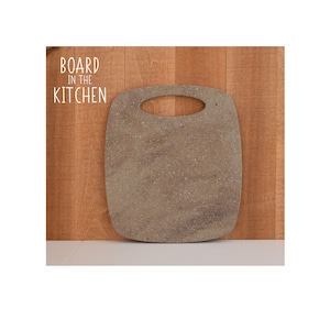 May include: A square-shaped cutting board with a speckled brown and gray surface. The board has a handle with a hole in the center. The board is sitting on a wooden surface.