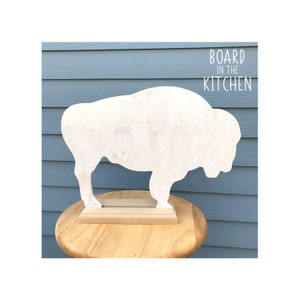 May include: White buffalo-shaped cutting board with a wood base. The board is on a wooden table in front of a blue wall. The text "BOARD in the KITCHEN" is printed on the wall.
