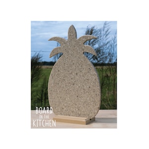 May include: A beige pineapple-shaped cutting board with a wood stand. The board is made of a speckled material and has a cutout design of a pineapple. The text "BOARD IN THE KITCHEN" is printed below the board.