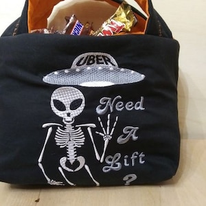 May include: Black Halloween trick or treat bag with an embroidered skeleton holding a hand up and saying "Need a Lift?" with a UFO above the skeleton that says "Uber".