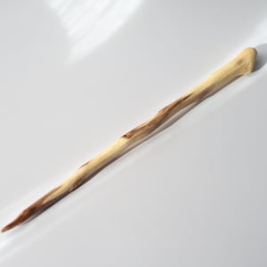 Könnte beinhalten: Ein handgefertigter Haarstab aus Holz mit einer natürlichen, hellen Holzmaserung und dunkelbraunen Streifen. Der Stab hat eine längliche, leicht unregelmäßige und spitz zulaufende Form mit einem abgerundeten, dickeren Ende und einer Spitze.