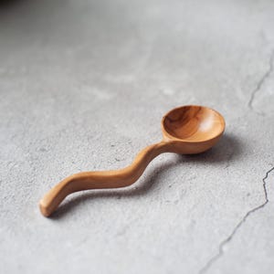 Könnte beinhalten: Ein kleiner, handgefertigter Holzlöffel mit gebogenem Griff und rundem Löffel. Der Löffel ist aus hellbraunem Holz gefertigt und hat ein natürliches, organisches Aussehen. Der Löffel wird wahrscheinlich zum Servieren oder Essen verwendet.